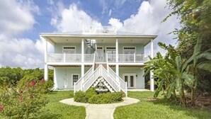 A two-story white house with a central staircase, surrounded by lush greenery and a clear sky.