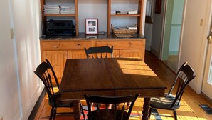 Een eethoek met een houten tafel en stoelen, een boekenkast met diverse spullen, en een klein model van een schip erboven.