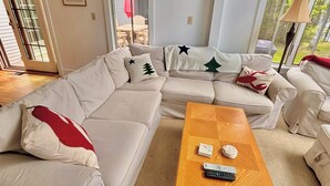 Een woonkamer met een beige hoekbank, een houten salontafel en een uitzicht op het groen door grote ramen.