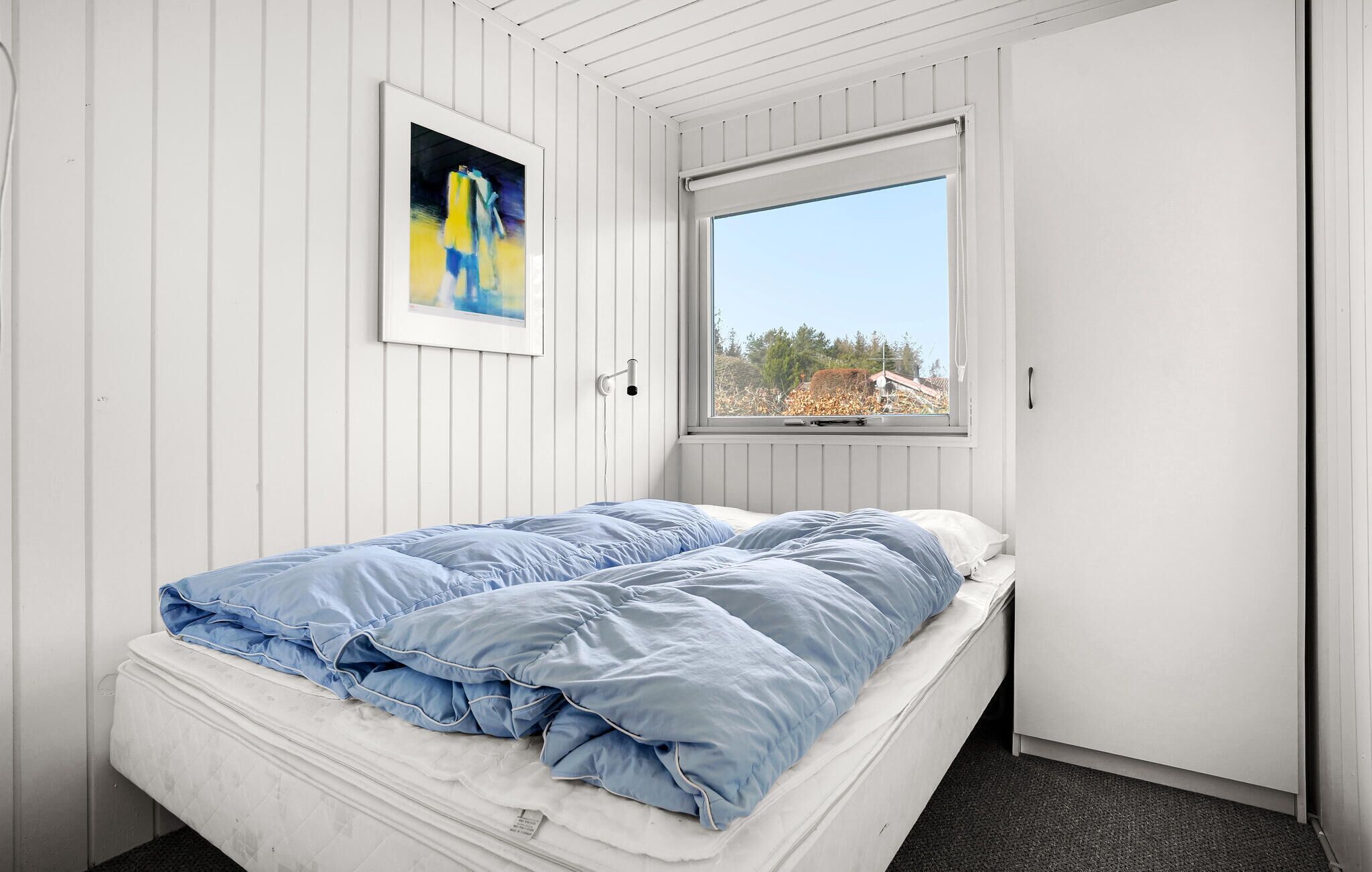 Um quarto pequeno, com painéis brancos, uma cama de solteiro, um quadro emoldurado e uma janela com vista para árvores e um edifício.