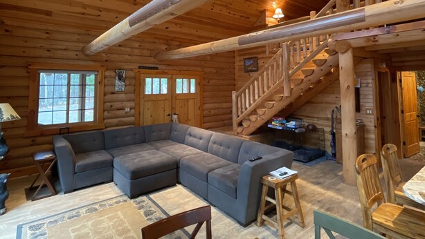 Interno di una baita di legno con un ampio divano componibile, un tavolo da pranzo con sedie, una scala e vista all'esterno attraverso una finestra.