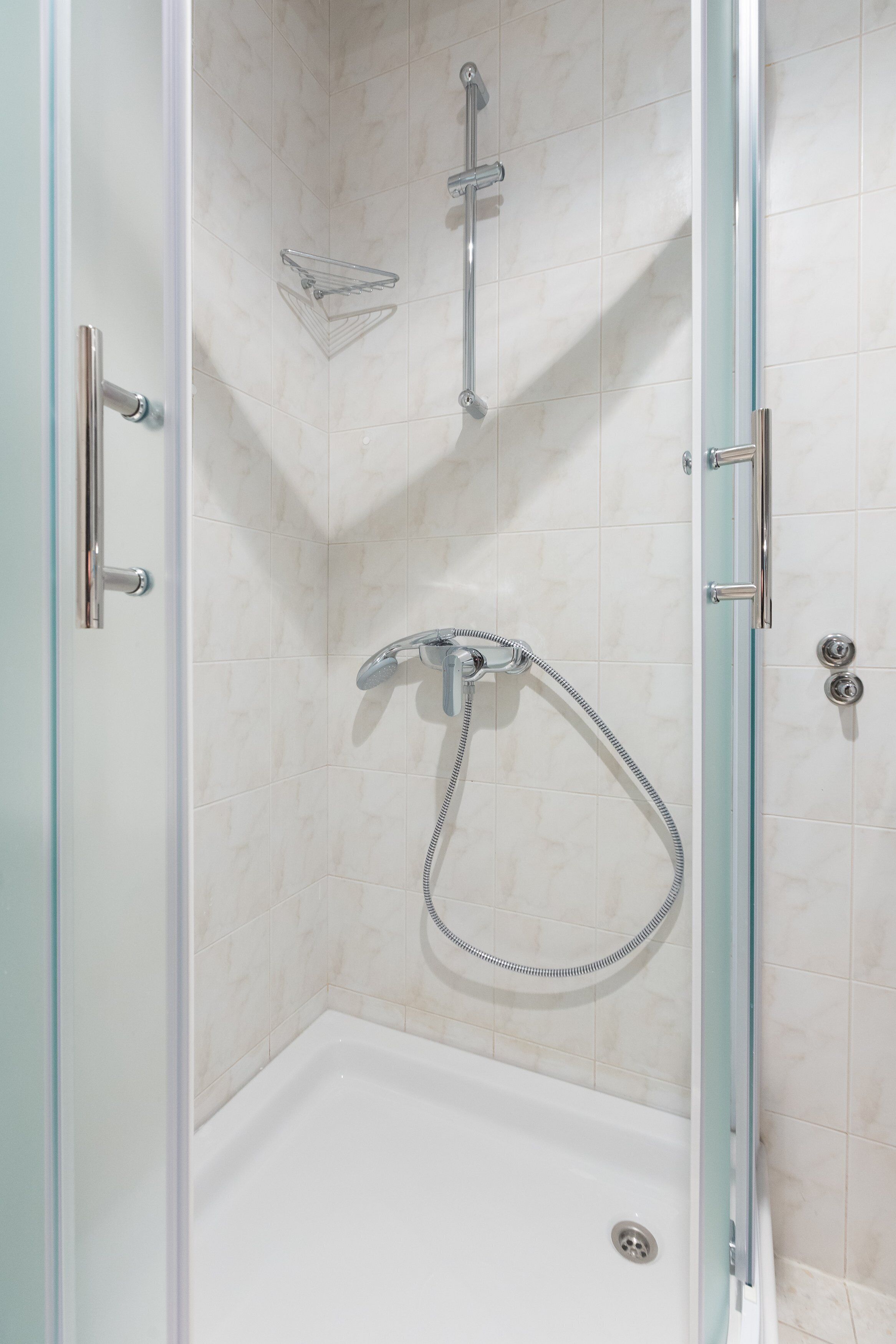 A glass-enclosed shower stall with a handheld showerhead, a glass shelf, and a door with a metallic handle.