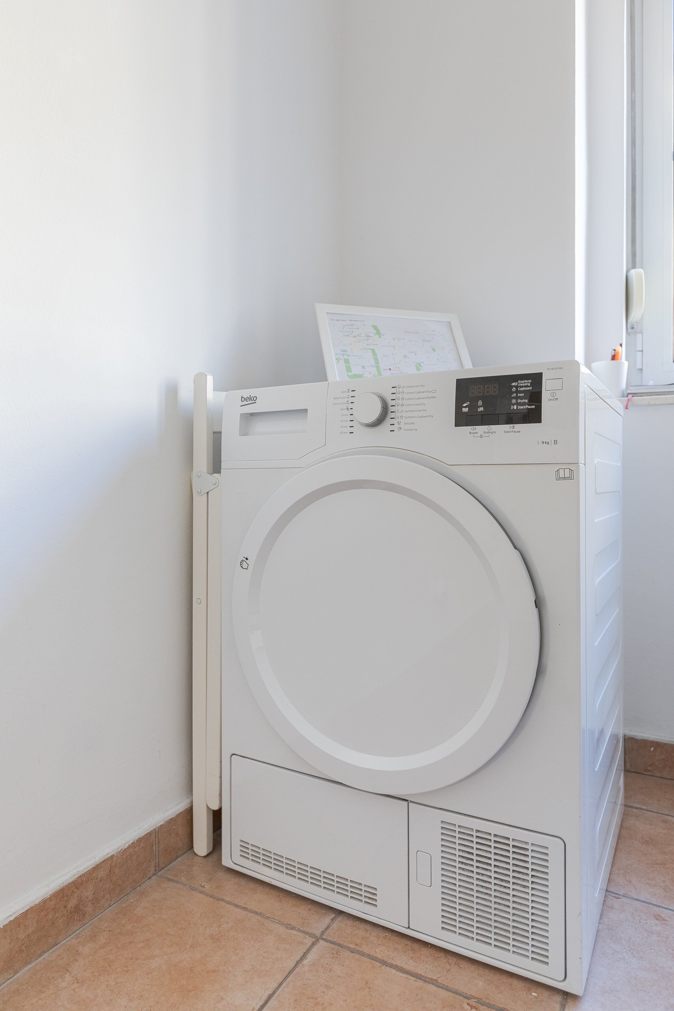 A front-loading washing machine with a digital display, located in a corner of a room with tiled flooring and a white wall.