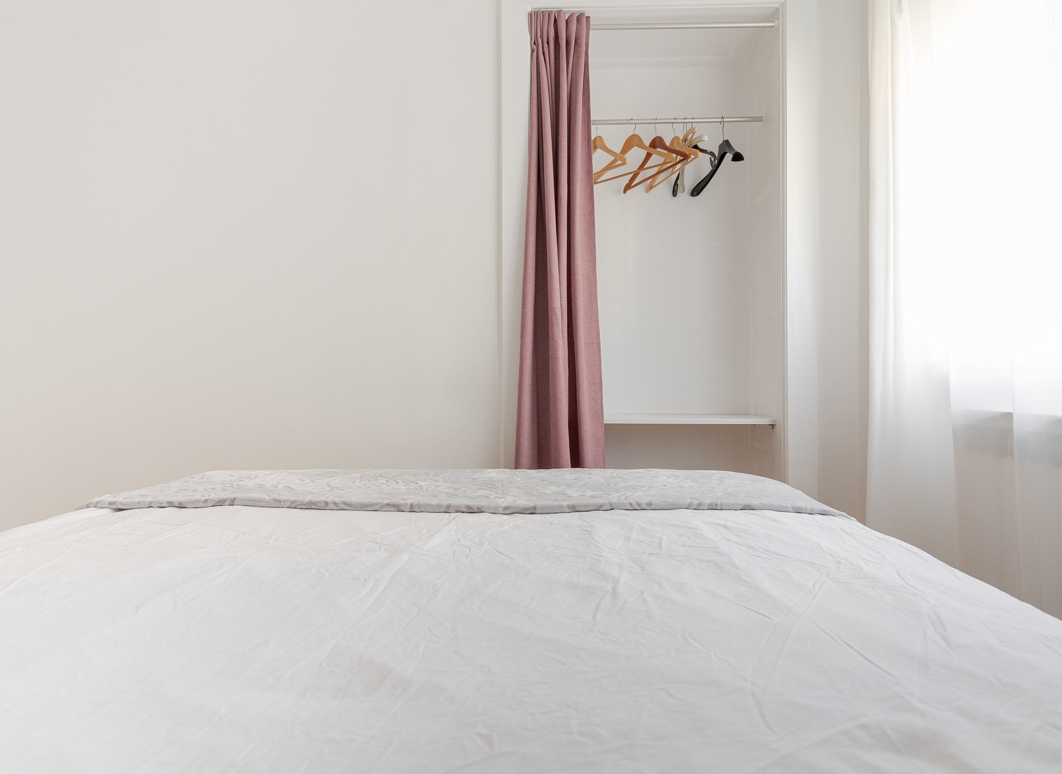A neatly made bed with a white comforter and a gray blanket, a pink curtain partially drawn, and a closet with hangers and a shelf.