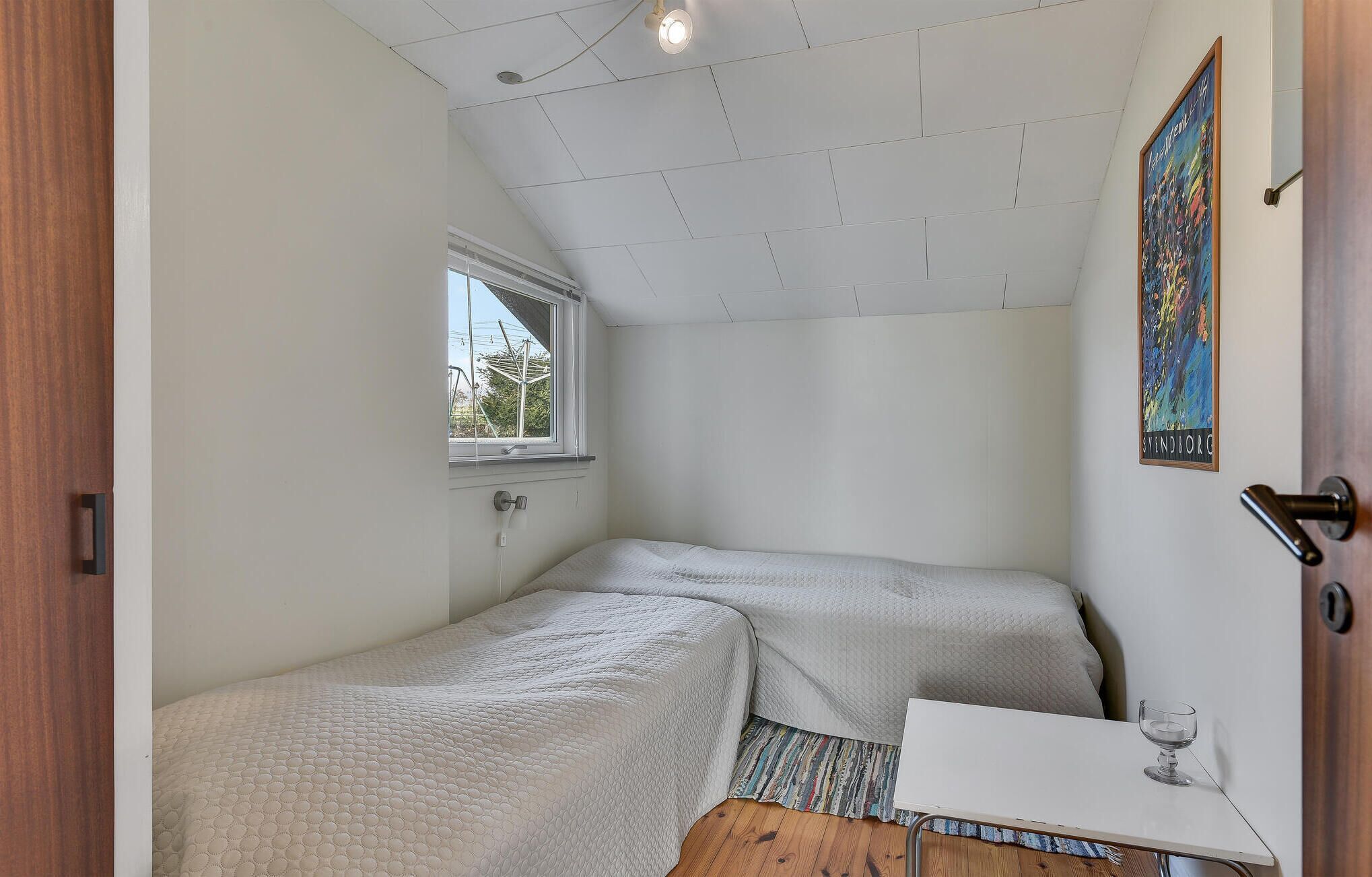 Una habitación pequeña y funcional con dos camas individuales, una mesita, una ventana y una puerta.