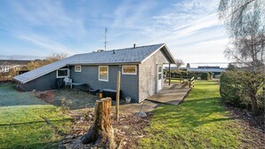 Ein Bungalow mit geneigtem Dach, einer kleinen Veranda und einem Garten mit einer Holzterrasse und einem Baumstumpf.