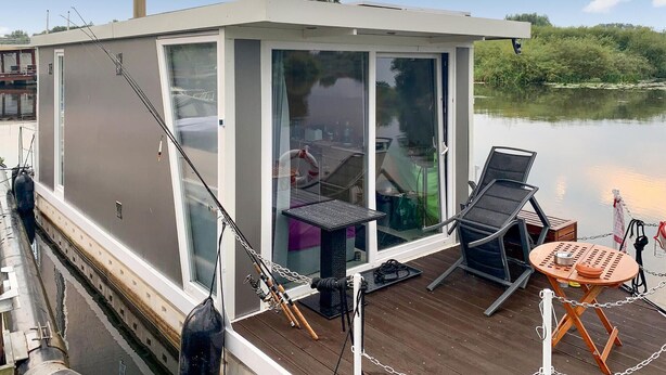 Une structure flottante moderne dotée d’une enceinte en verre, d’une terrasse avec des chaises et une table, ainsi que de matériel de pêche.