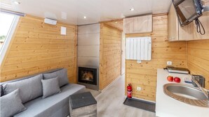 Une cabane compacte et moderne, avec un intérieur en bois, un canapé, une cheminée, un coin cuisine équipé d’un évier et un téléviseur.