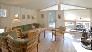 Ein gemütliches Zimmer mit Holzboden, einem grünen Sofa, einem Holztisch und einem Sessel. An den Wänden hängen Bilderrahmen und es gibt ein Fenster mit Jalousien.