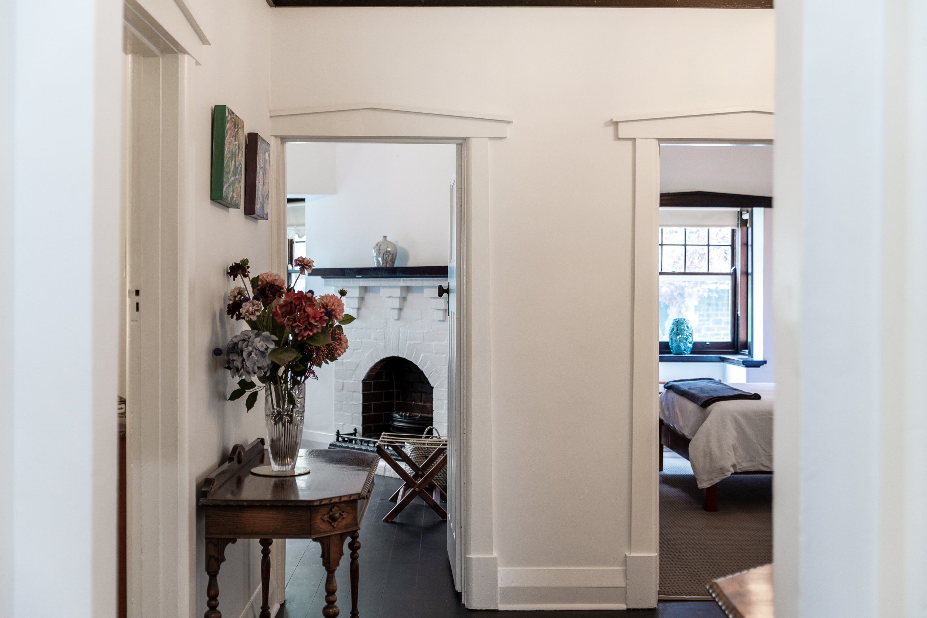 Un couloir avec une cheminée en briques blanches, une table à manger ornée d’un vase de fleurs et un lit dans la pièce adjacente.