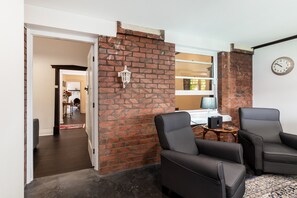 A room with a brick wall, two black leather chairs, a clock, and a small table with a lamp.