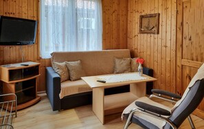 Una habitación acogedora con detalles de madera, un sofá, una mesita, una silla, un televisor y una ventana con cortinas translúcidas.