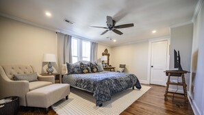Quarto com cama, uma cadeira, abajur, ventoinha no teto e um suporte de madeira.