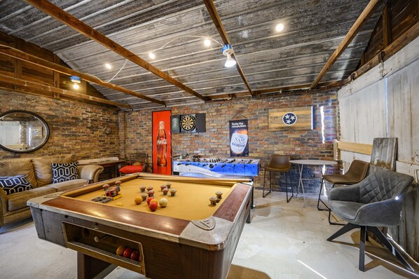 A pool table with balls, a foosball table, and a dartboard in a room with brick walls and wooden beams.