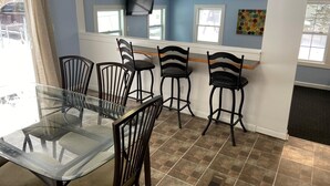 Un comedor con mesa de vidrio y sillas de madera, televisor montado en la pared y ventana con vistas al exterior.