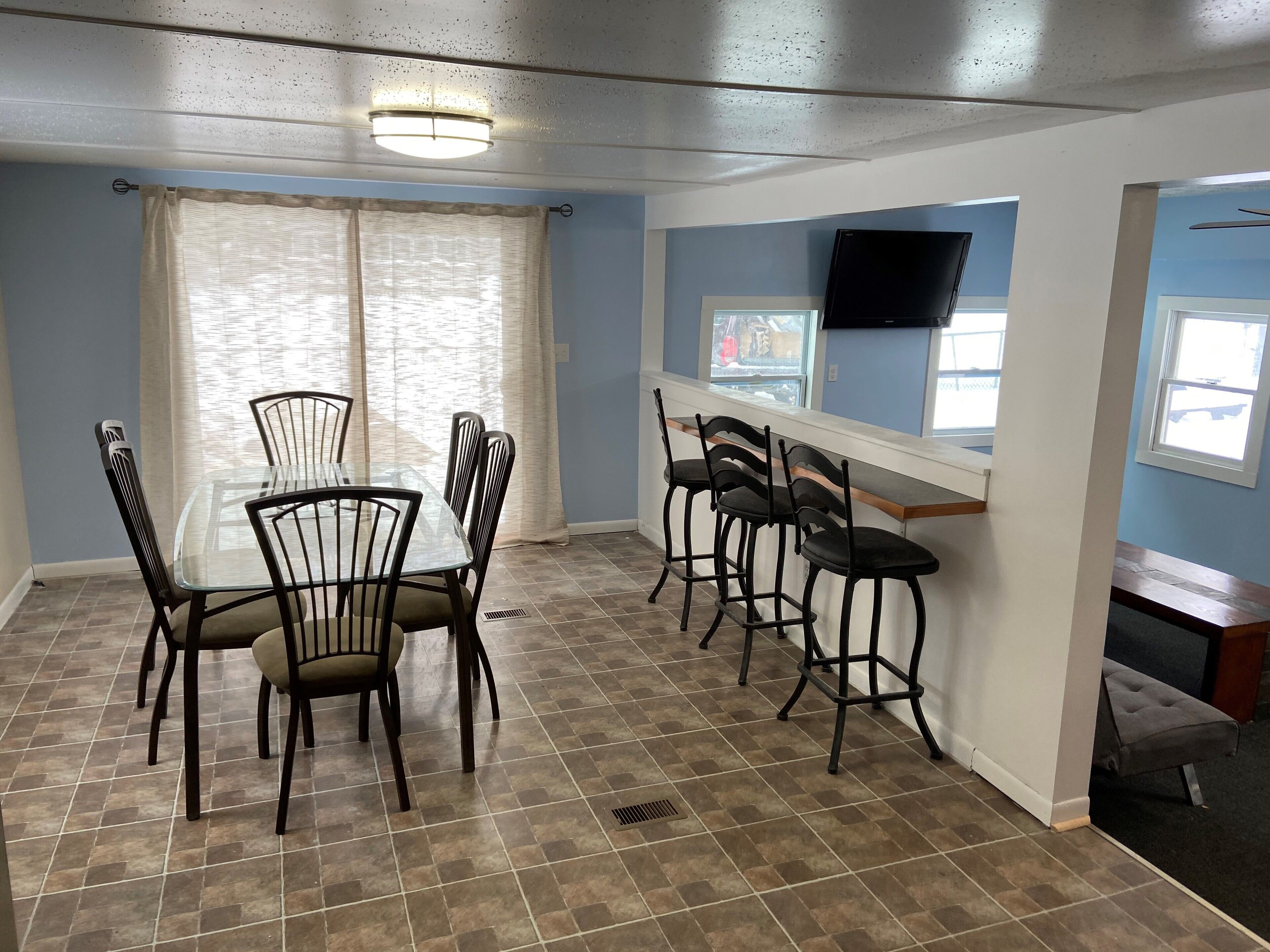 A dining area with a glass table and chairs, a bar counter with stools, and a TV mounted on the wall.