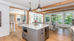 A spacious kitchen with a large island, wooden beams, and large windows offering a view of greenery.