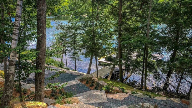 A serene lakeside garden with stone pathways, lush greenery, and a dock extending into the water.