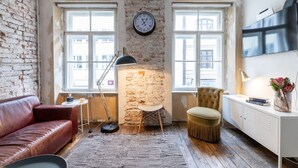 A room with exposed brick walls, a leather sofa, a round table with a lamp, a clock on the wall, and a TV on the wall. - Loft Munich Old Town (München)
