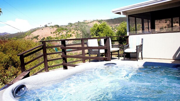 A hot tub with a wooden deck and chairs, overlooking a hilly landscape.