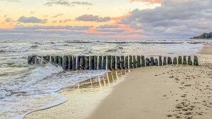 Een strand met golven die tegen een houten golfbreker slaan, een zandstrand en een bewolkte lucht.