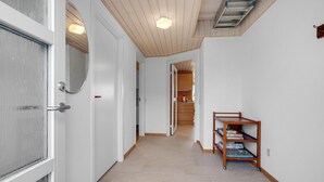 Ein Flur mit weißen Wänden, einer Holzbalkendecke und einem gefliesten Boden. Links eine Tür, rechts ein Regal mit Büchern, und am Ende eine Küchenzeile.