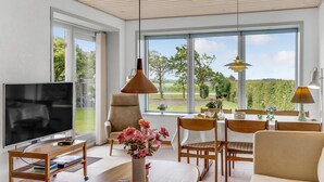 Ein modernes Wohnzimmer mit Fernseher, bequemen Sitzgelegenheiten, einem Essbereich und großen Fenstern mit Blick auf Grünflächen.