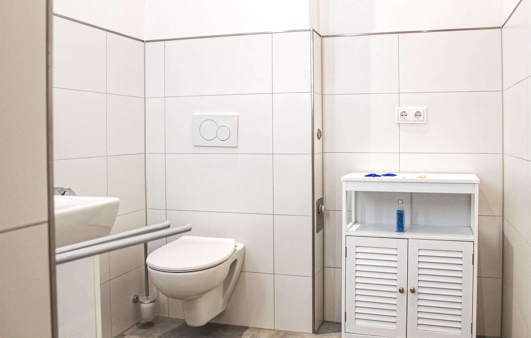 Pieni, valkoinen kylpyhuone, jossa on wc-istuin, kaide ja yläosassa pullo.