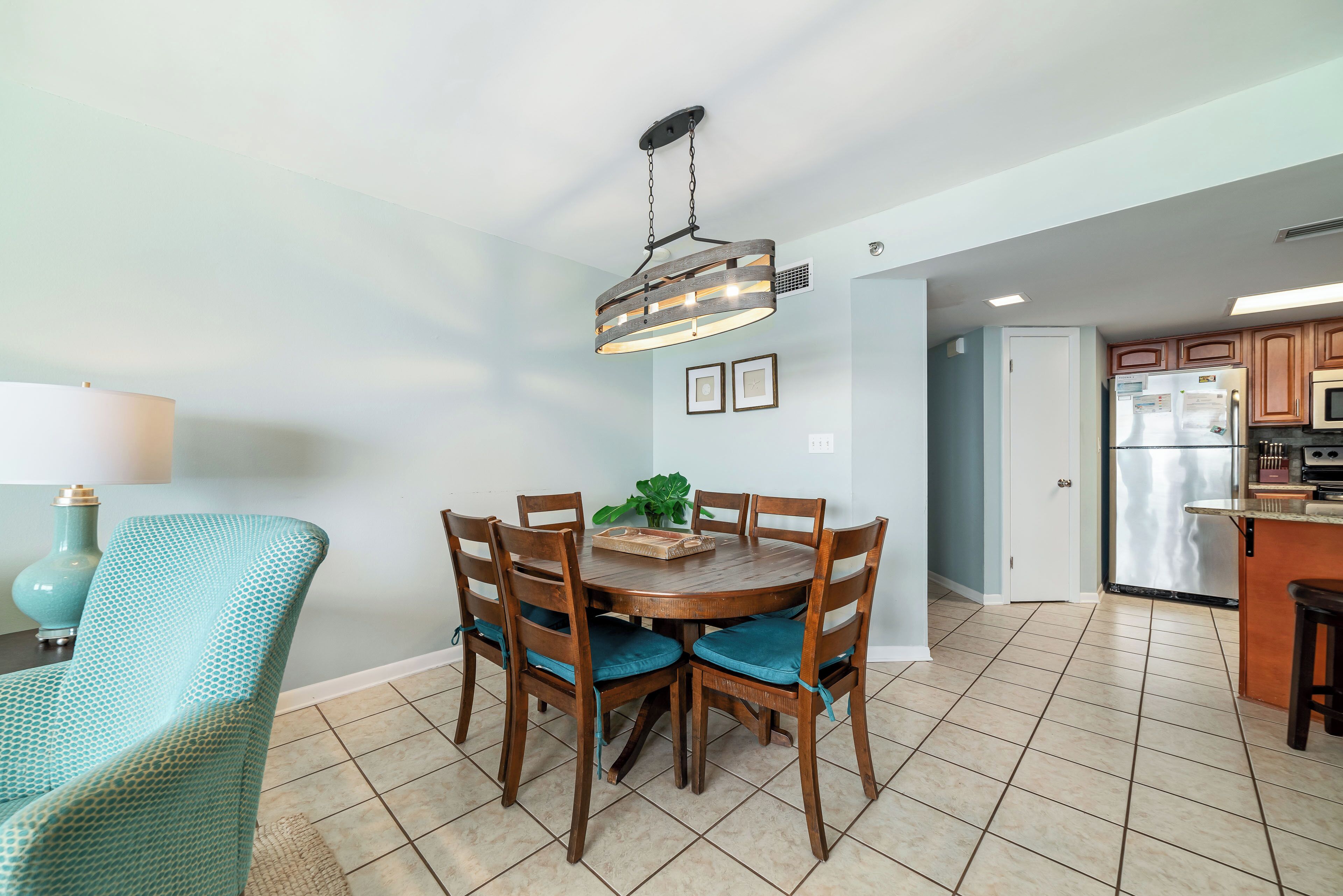 A dining area with a wooden table and chairs, a hanging light fixture, and a kitchen in the background.