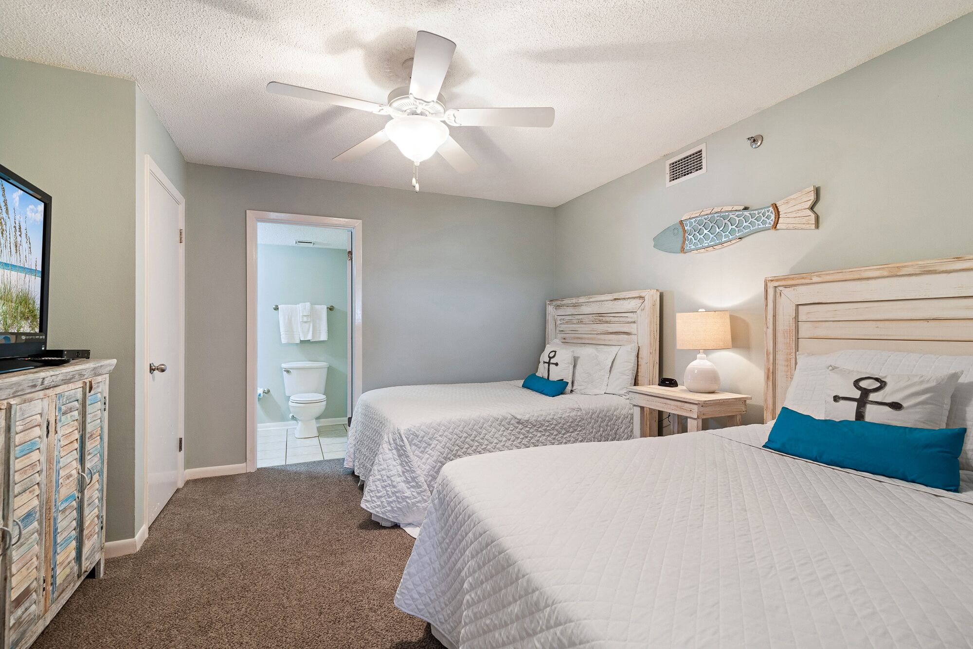 A bedroom with two beds, a ceiling fan, a bathroom with a toilet and towel rack, and a television on a cabinet.