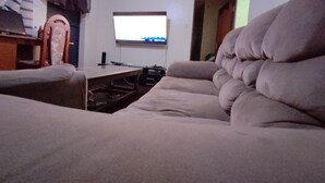 Un salon avec un canapé beige, un téléviseur fixé au mur et une table en bois avec une chaise.