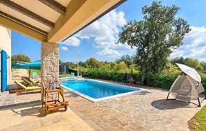 A swimming pool with lounge chairs and a sun umbrella, surrounded by stone tiles and greenery. - Stunning home in Naselje Muzini with sauna (Naselje Muzini)