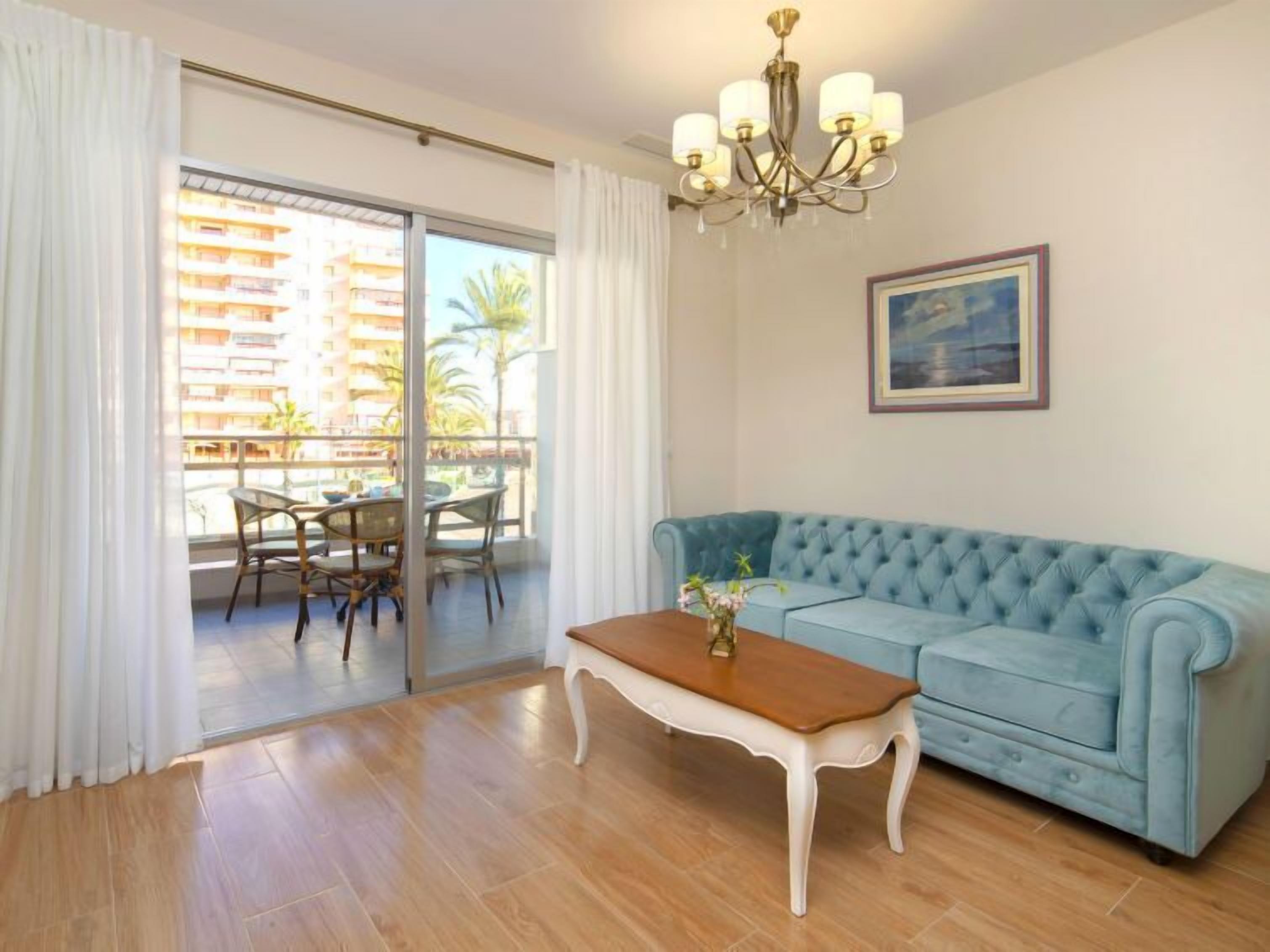 Una sala de estar con un sofá azul claro, una mesa de centro de madera y una araña. Hay un balcón con una mesa y sillas, y un cuadro en la pared.