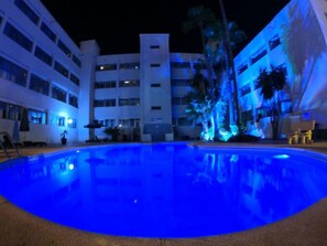 A nighttime view of a hotel pool area with a blue-lit pool, palm trees, and a multi-story building in the background.