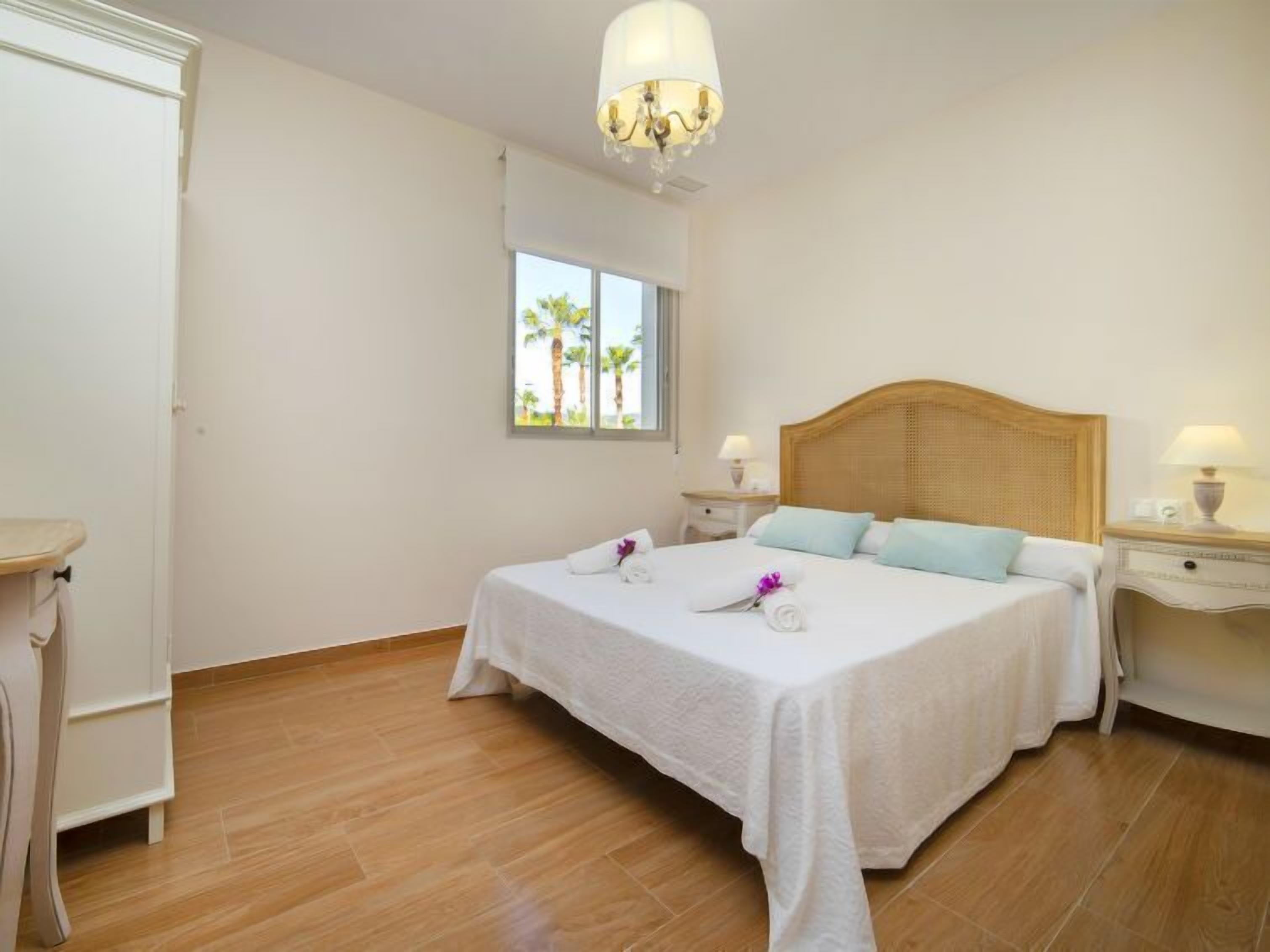 Habitación de hotel con cama, mesitas de noche, ventana con vista a palmeras y un candelabro.