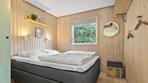 A modern bedroom with a wooden paneled wall, a large window with a view of greenery, a bedside table with a lamp, and a small wicker chest.