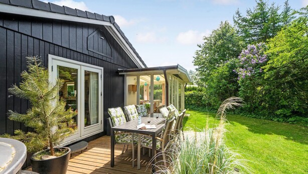 A wooden deck with a table and chairs, a covered seating area, and a garden with various plants and trees.