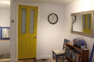 Una habitación con una puerta amarilla, un reloj, un espejo y una estantería con libros y una bolsa azul.