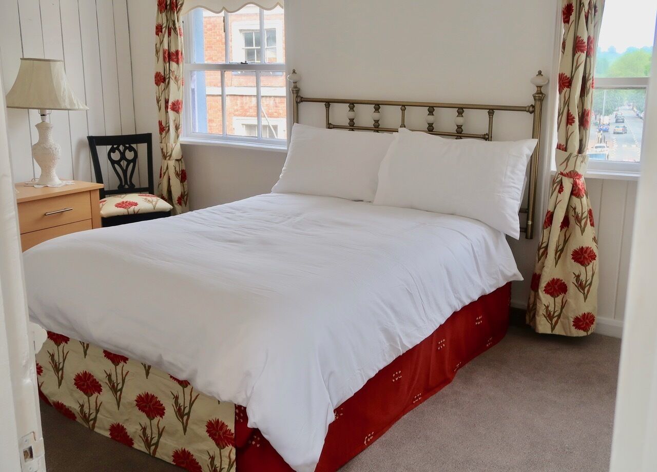 Una cama bien hecha con sábanas blancas y falda roja, una mesita de noche de madera con una lámpara y cortinas florales.