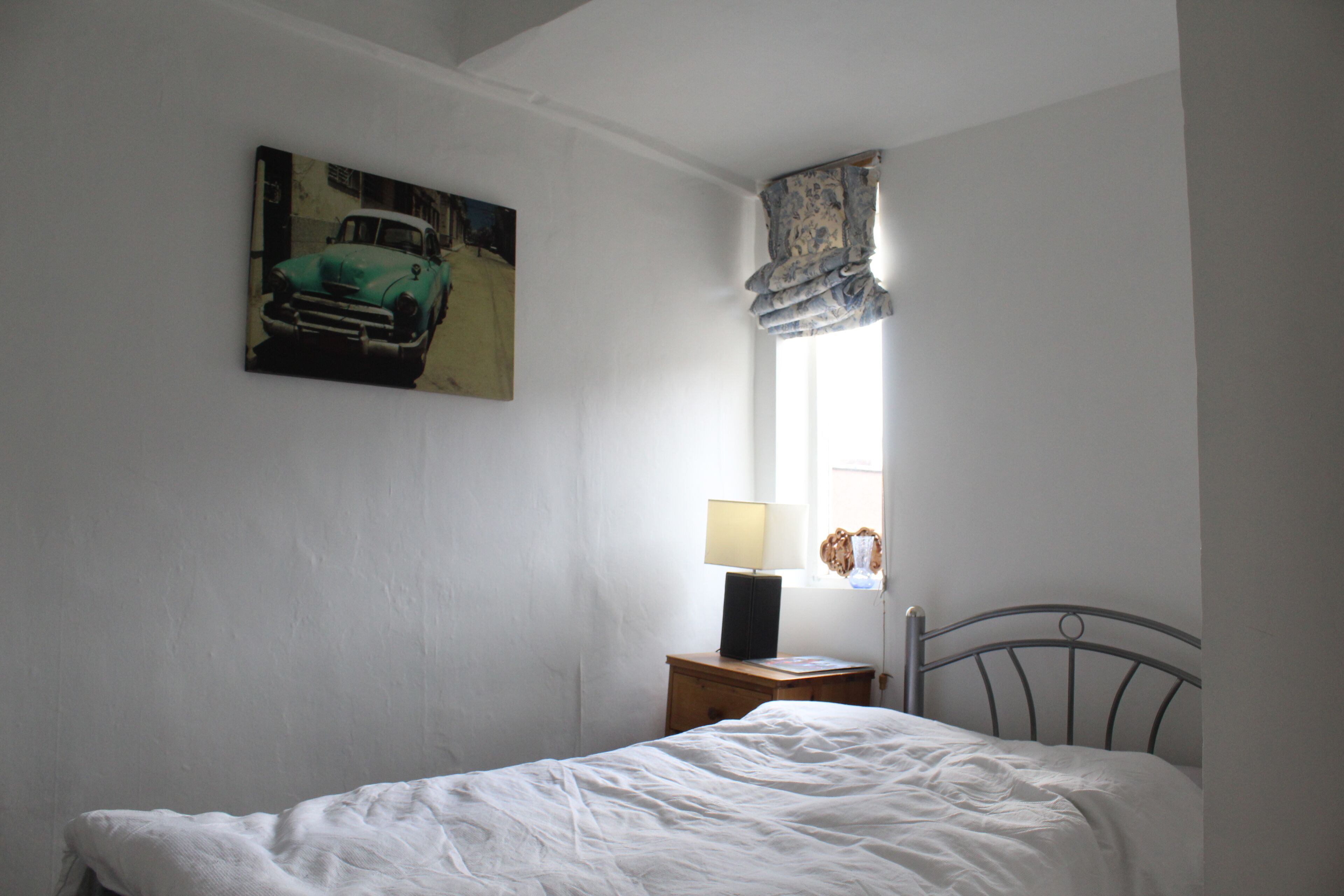 A bedroom with a bed, a nightstand with a lamp, a painting on the wall, and a window with a curtain.
