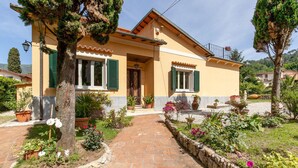 Una casa de dos pisos con entrada pavimentada, contraventanas verdes y un jardín con diversas plantas y flores.