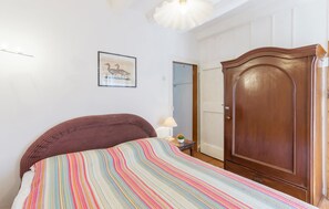 A bedroom with a striped bedspread, a wooden wardrobe, a framed picture on the wall, and a lamp on a bedside table. - Nice home in Funtana with WiFi (Funtana)