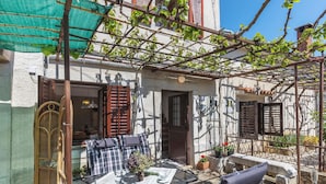 Una zona d'estar exterior acollidora amb taula i cadires, una pèrgola amb plantes enfilades i una porta que dona a l'interior.