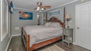 Une chambre à coucher avec un lit, un ventilateur de plafond, une table de chevet, une lampe, un tableau et une pancarte sur laquelle on peut lire « Beach House ».