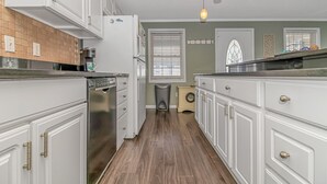 Une cuisine avec des meubles blancs, un lave-vaisselle, un lave-linge et une fenêtre avec des stores.
