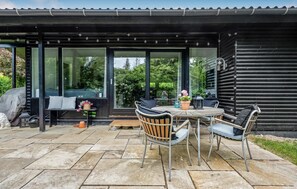 An outdoor patio area with a round table and chairs, a covered seating area with cushions, and a view of the surrounding greenery.