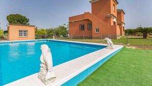 A swimming pool with two stone sculptures, a large house with a red facade, and a well-maintained lawn.