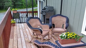 Une terrasse en bois, meublée de meubles en osier, avec un barbecue et des plantes en pot.