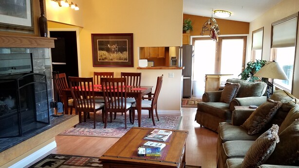 Un salon avec une cheminée, des meubles en bois, un coin repas et une cuisine à l’arrière-plan.