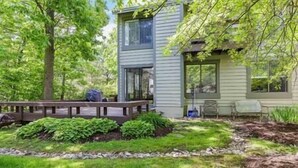 A two-story house with a deck, surrounded by lush greenery and trees.